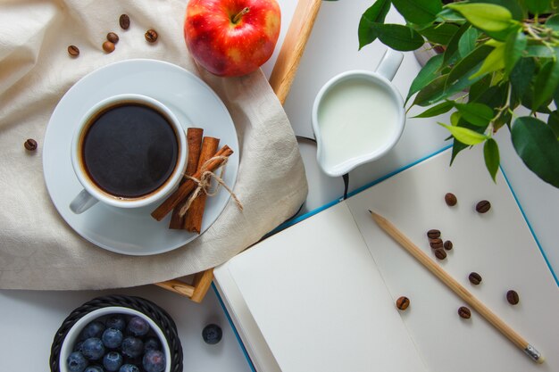 Vista superior de una taza de café con leche, manzana, canela seca, arándanos, planta, lápiz y cuaderno sobre superficie blanca. horizontal