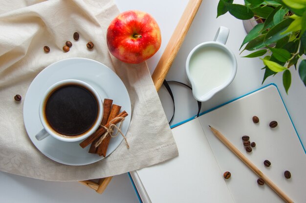 Vista superior de una taza de café con leche, manzana, canela seca, planta, lápiz y cuaderno sobre superficie blanca. horizontal