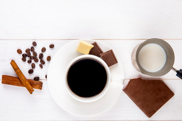 Vista superior de una taza de café con palitos de canela, barra de chocolate y granos de café esparcidos sobre fondo blanco de madera