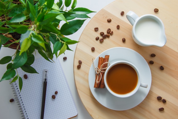 Vista superior de una taza de café con plantas, granos de café, leche, canela seca, cuaderno y bolígrafo en la plataforma y superficie blanca. horizontal