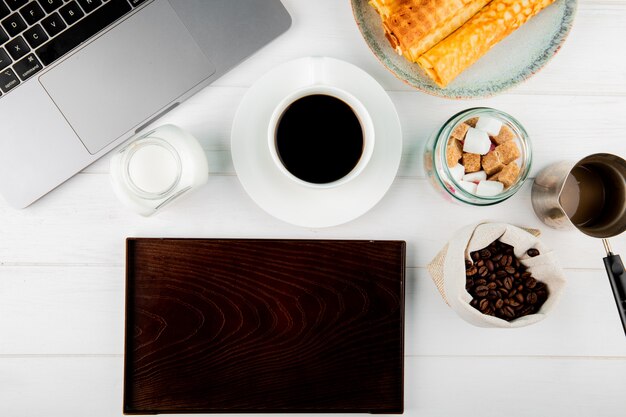 Vista superior de una taza de café con rollos de oblea granos de café en un saco portátil y una tabla de madera sobre fondo blanco.