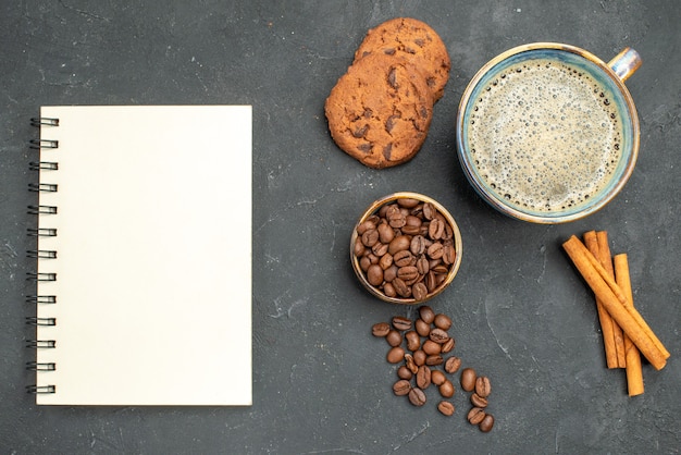 Vista superior de una taza de café con semillas de café, canela, galletas, un notebok sobre fondo oscuro aislado