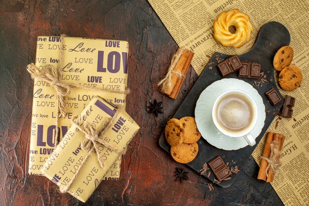 Vista superior de la taza de café en la tabla de cortar de madera en un periódico antiguo galletas canela limas barras de chocolate hermosas cajas de regalo sobre fondo oscuro