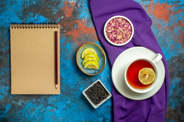 Vista superior de una taza de cuencos de té con pétalos de flores secas y rodajas de té de limón mantel morado apencil en el cuaderno sobre la superficie azul roja