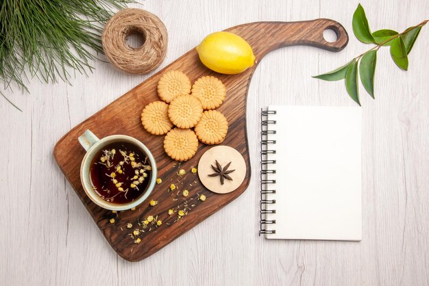 Vista superior de una taza de galletas de té, limón, anís estrellado, una taza de té de hierbas en la tabla de madera, hojas junto al cuaderno blanco y ramas de árboles de Navidad.