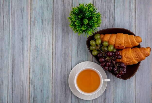 Vista superior de la taza de hot toddy y croissants uvas en un recipiente con planta sobre fondo de madera con espacio de copia