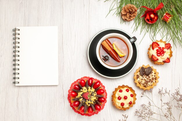 Vista superior de una taza de tartas de pastel de bayas de té de canela y limón y hojas de pino con juguetes navideños y un cuaderno en el suelo de madera blanca