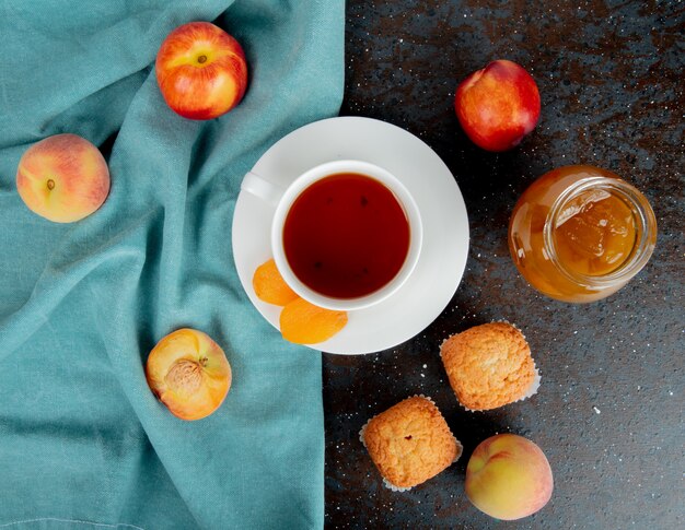 Vista superior de una taza de té con albaricoques secos y duraznos maduros frescos sobre tela azul y magdalenas con un tarro de mermelada de durazno en negro