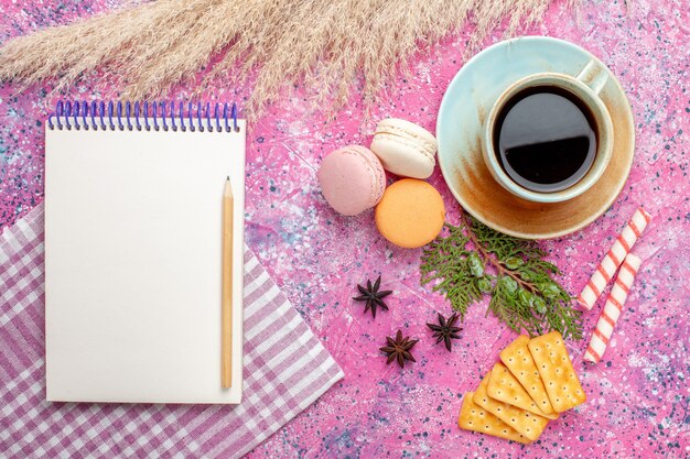 Vista superior de una taza de té con bloc de notas de galletas y macarons en superficie rosa