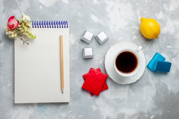 Vista superior de la taza de té caliente dentro de la taza blanca con el bloc de notas de caramelos de chocolate y paquete plateado en el escritorio de luz, beber la hora del té dulce