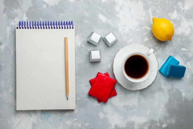 Vista superior de la taza de té caliente dentro de la taza blanca con el bloc de notas de caramelos de chocolate y paquete plateado en el escritorio de luz, beber la hora del té de galletas dulces