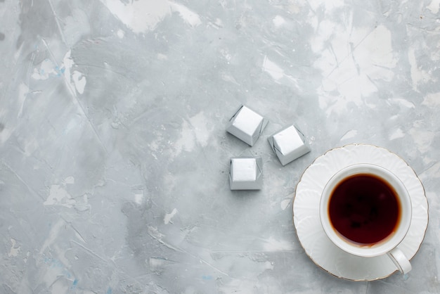 Vista superior de la taza de té caliente dentro de la taza blanca en un plato de vidrio con caramelos de chocolate de paquete plateado en el escritorio de luz, bebe galleta de chocolate dulce