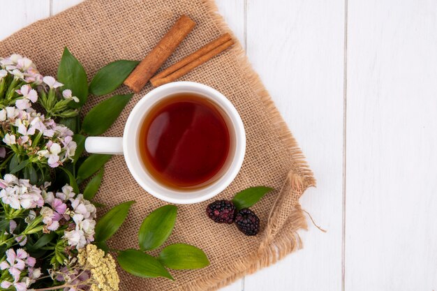 Vista superior de la taza de té con canela y flores en una servilleta beige sobre una superficie blanca