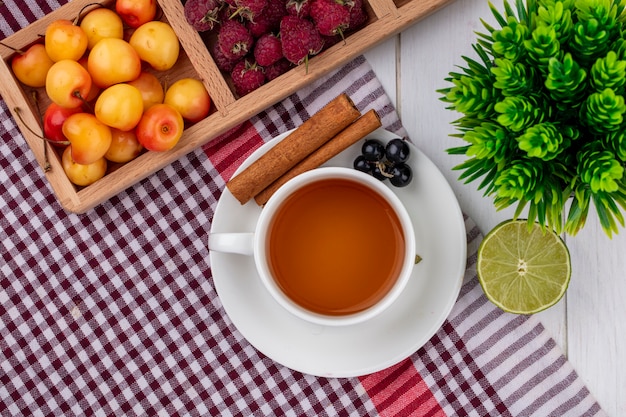 Vista superior de la taza de té con canela, grosella, frambuesas y cerezas blancas sobre una toalla a cuadros rojos