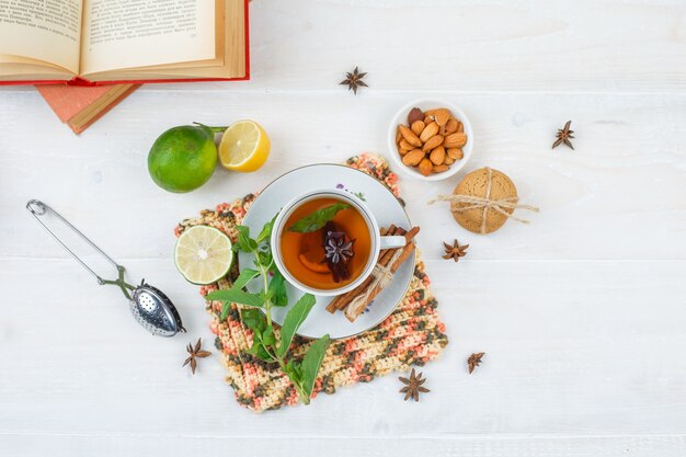Vista superior de la taza de té con canela y limón en un mantel cuadrado con limas, un tazón de almendras, colador de té y libros sobre superficie blanca