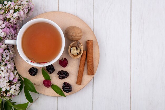 Vista superior de la taza de té con canela, nueces, frambuesas y moras en un soporte con flores sobre una superficie blanca