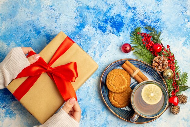 Vista superior de la taza de té canela palitos de galletas en el platillo de Navidad presente en mano femenina en el cuadro azul