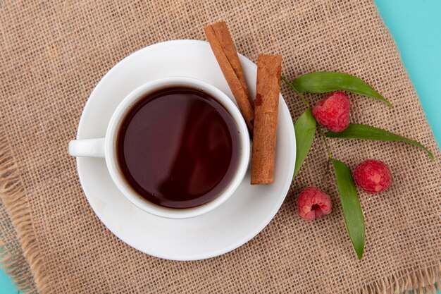 Vista superior de la taza de té y canela en platillo con frambuesas y hojas de cilicio