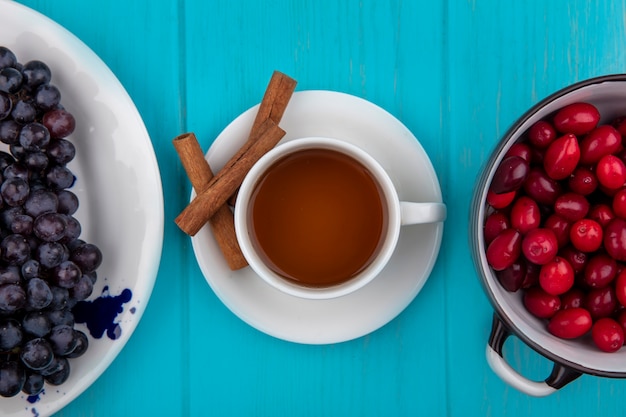 Vista superior de la taza de té con canela en el platillo y el plato de uva tazón de fuente de bayas de cornel sobre fondo azul.