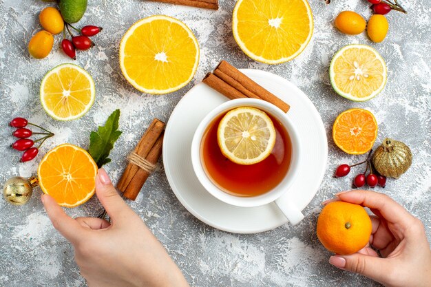 Vista superior de una taza de té cortar limones canela en rama mandarina y cortar limón en manos femeninas sobre fondo gris