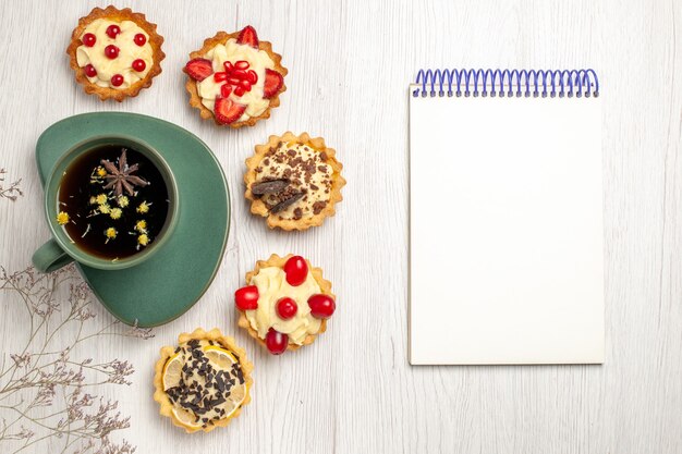 Vista superior de una taza de té y diferentes galletas a la izquierda y un cuaderno en el lado derecho de la mesa de madera blanca.