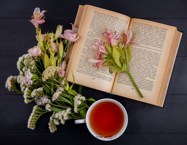 Vista superior de la taza de té con flores de color rosa claro y un libro abierto sobre una superficie negra