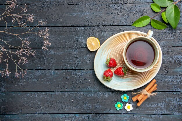 Vista superior de una taza de té y fresas en platillo rodaja de limón flor de canela dulces hojas sobre la mesa de madera oscura.