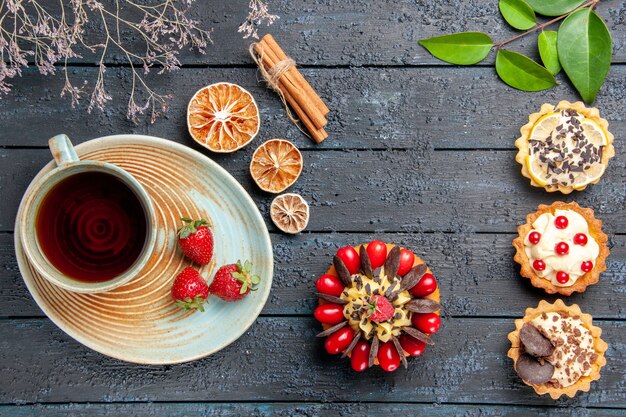 Vista superior de una taza de té y fresas en un platillo, tartas de naranjas secas, hojas y tarta de bayas en la mesa de madera oscura.