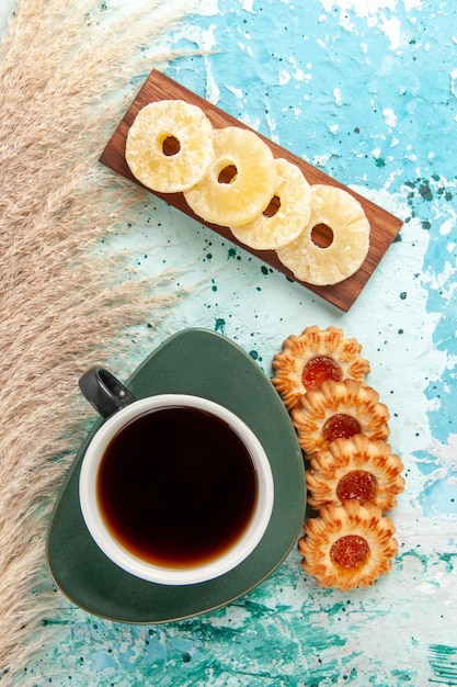 Vista superior de una taza de té con galletas y anillos de piña secos sobre una superficie azul claro