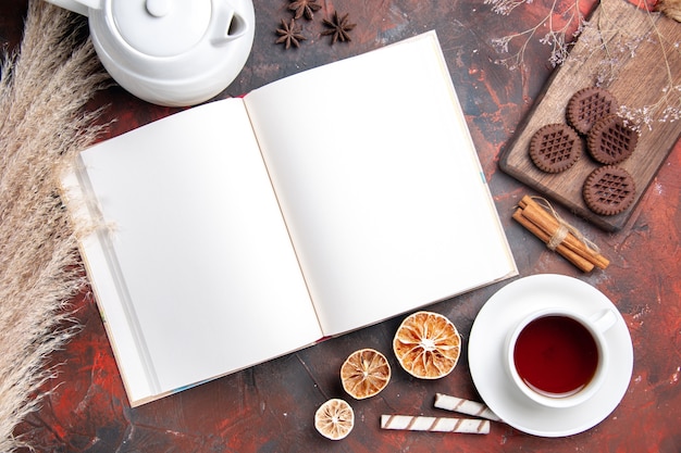 Vista superior de la taza de té con galletas y cuaderno sobre mesa oscura