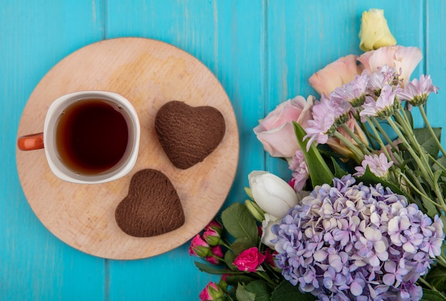 Vista superior de la taza de té y galletas en forma de corazón en la tabla de cortar con flores sobre fondo azul.