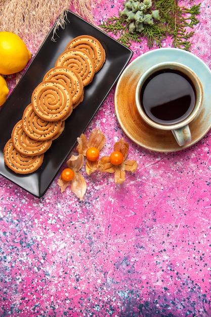 Vista superior de la taza de té con galletas y limón en el escritorio de color rosa claro.