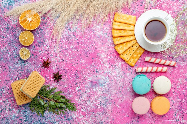 Vista superior de una taza de té con galletas y macarons franceses en el escritorio de color rosa claro