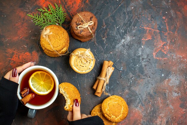 Vista superior de una taza de té y galletas en la mano femenina, palitos de canela, galletas atadas con una cuerda en el espacio libre de la mesa de color rojo oscuro