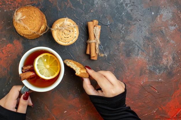 Vista superior de una taza de té y galletas en la mano femenina, palitos de canela, galletas atadas con una cuerda en el espacio libre de la mesa de color rojo oscuro