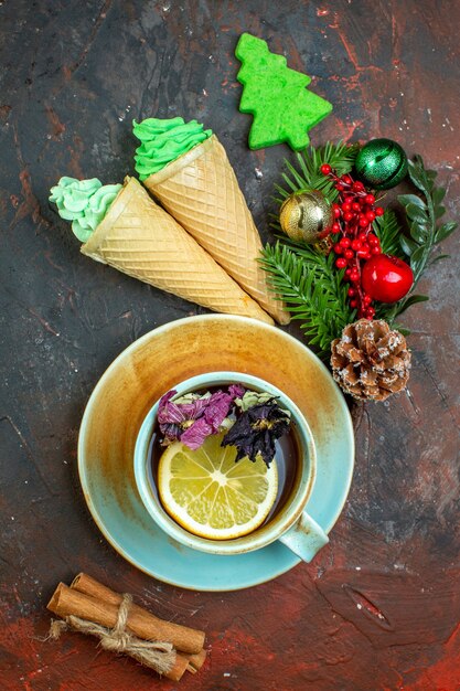 Vista superior de la taza de té, helados, ramas de canela, rama de árbol de Navidad con juguetes en la mesa de color rojo oscuro