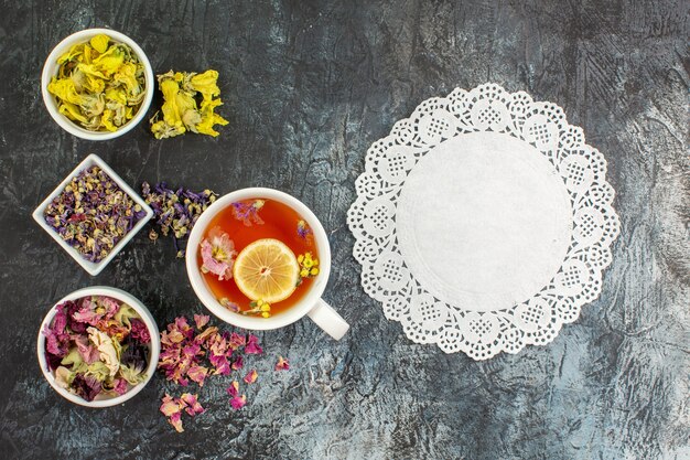 Vista superior de una taza de té de hierbas con cuencos de flores secas y encaje sobre fondo gris