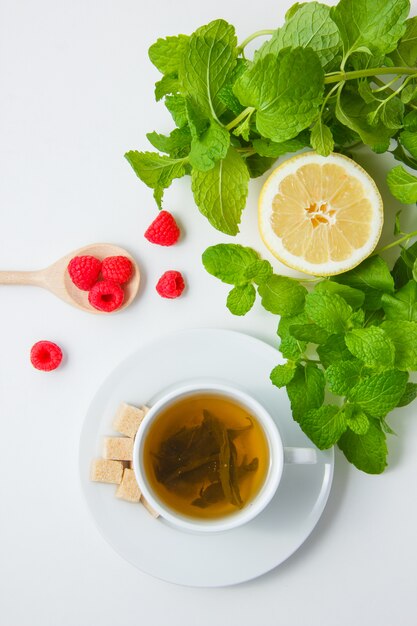 Vista superior de una taza de té con limón, azúcar, hojas de menta, frambuesa sobre superficie blanca. vertical