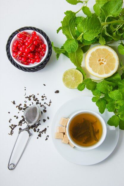 Vista superior de una taza de té con limón, azúcar, hojas de menta, frambuesa sobre superficie blanca. vertical