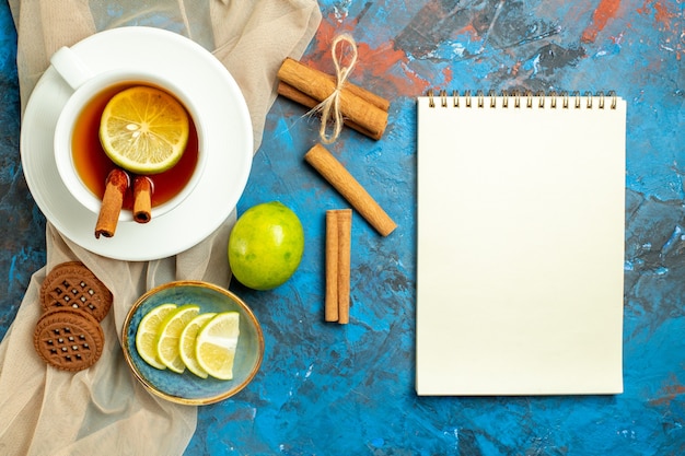 Vista superior de una taza de té con limón y canela, galletas de mantón beige, cuaderno de limón sobre superficie roja azul