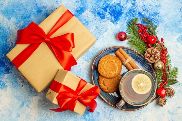 Vista superior de la taza de té con limón, canela, galletas en platillo, regalos de Navidad en la mesa azul