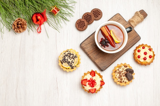 Vista superior de una taza de té de limón y canela en la tabla de cortar tartas galletas y hojas de pino con juguetes de Navidad en el suelo de madera blanca