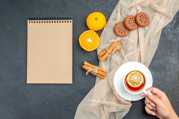 Vista superior de una taza de té con limón en mano femenina canela palitos de galletas en el bloc de notas de color beige mantón naranja sobre superficie oscura