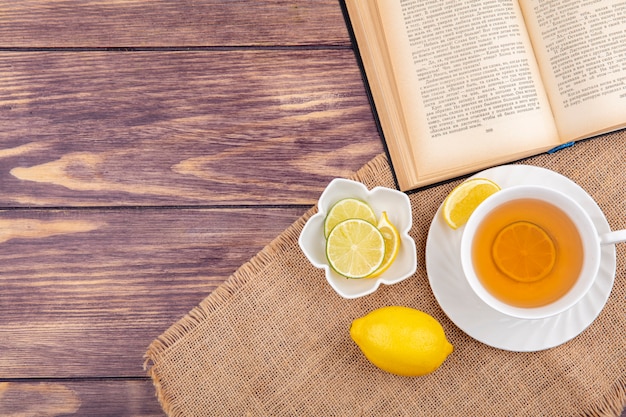 Vista superior de una taza de té con limones frescos en un tazón blanco sobre tela de saco en madera con espacio de copia