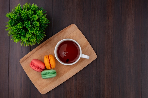 Vista superior de la taza de té con macarons de colores sobre una tabla de cortar sobre una superficie de madera