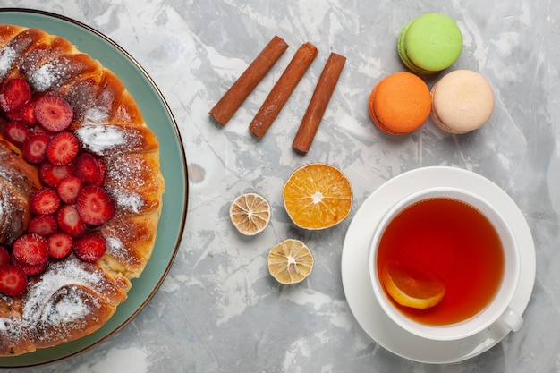 Vista superior de la taza de té con macarons franceses y tarta de fresa sobre superficie blanca