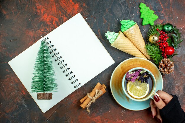 Vista superior de la taza de té en la mano femenina mini bloc de notas de helados de árbol de Navidad en la mesa de color rojo oscuro