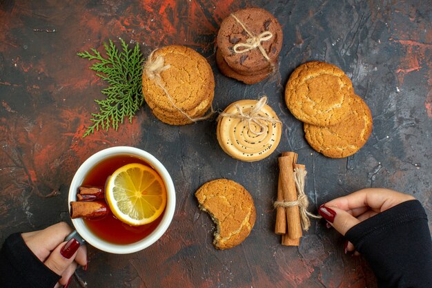 Vista superior de la taza de té en mano femenina palitos de canela diferentes galletas atadas con una cuerda en la mesa de color rojo oscuro