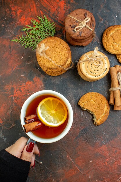 Vista superior de la taza de té en mano femenina palitos de canela diferentes galletas atadas con una cuerda en la mesa de color rojo oscuro