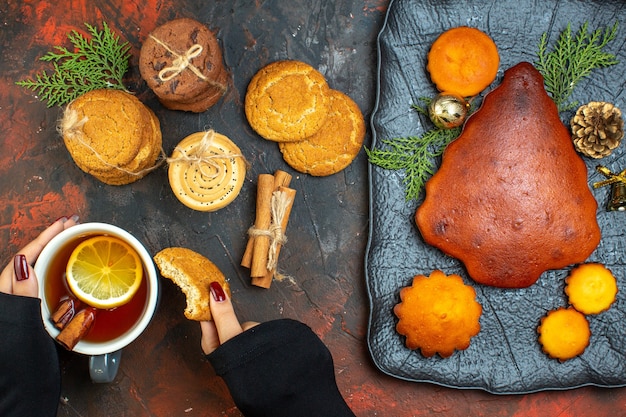 Vista superior de la taza de té en la mano femenina palitos de canela galletas atadas con pastel de cuerda en la placa en la mesa de color rojo oscuro